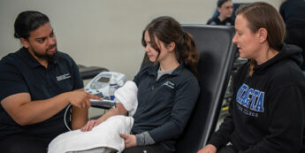 A student demonstrates a blood-pressure monitor on a seated student while another student watches.