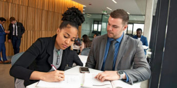 Black female student takes notes while white male student reads to her from textbook in brightly lit study area