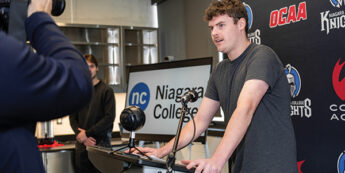 College student speaks at a podium during a press conference, with microphones, a photographer, and Niagara College Knights backdrop