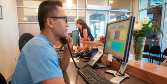 A male student is seated at a front desk, attending a call while using a computer.
