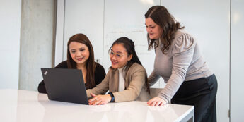 three female students are focused on a laptop, engaged in conversation while looking at the screen