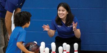 Female student engages a child to throw a bowling ball at bowling pins in the gym while a male student looks at the pair.