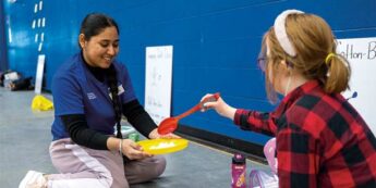 Female student interacting with child on the floor of a gym.