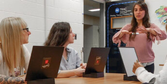 Professor speaking passionately to three attentive students seated at a table with laptops in a brightly lit room.