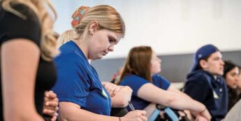 A group of students seated in a classroom. One female student in the foreground, is focused on writing notes.