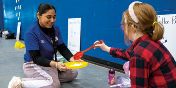 Female student interacting with child on the floor of a gym.