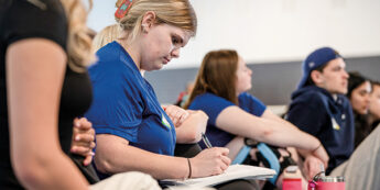 A group of students seated in a classroom. One female student in the foreground, is focused on writing notes.