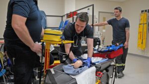 Paramedic student with a professor practising loading a newborn baby manikin on a stretcher while another student observes.