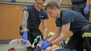 Two paramedic students practising on a hospital training manikin using a manual resuscitator while on the ground.