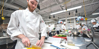 Male student in chef whites dices yellow pepper in an active kitchen laboratory classroom