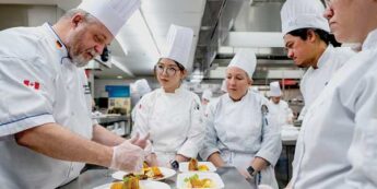Teaching chef in chef whites showing multiple student chefs in chef whites how to plate a dish in a teaching kitchen.