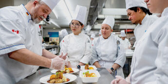 Teaching chef in chef whites showing multiple student chefs in chef whites how to plate a dish in a teaching kitchen.