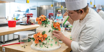 Female student in chef coat decorates a cake with orange flowers and green icing
