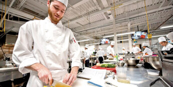 Male student in chef whites dices yellow pepper in an active kitchen laboratory classroom