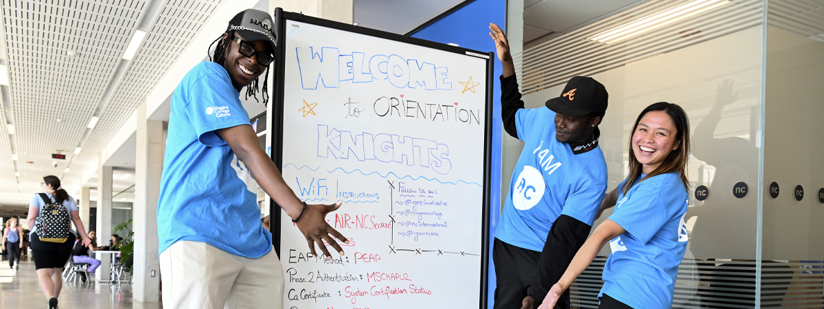 Three diverse students gesture at a white board covered in welcome messages