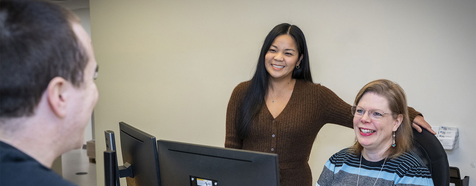 Two smiling female colleagues work at computer monitors in an office while a male coworker faces them