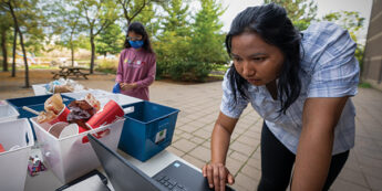Students sort items beside recycling bins in a park one types on a laptop