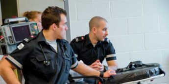 Two paramedics in uniform stand near a medical stretcher and equipment