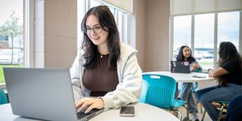 Student working on laptop with phone beside her at a table in a public area.