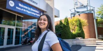 A smiling student stands outdoors at a campus main entrance