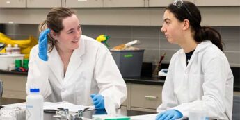 Two students in lab coats and blue gloves converse at a laboratory table with notebooks and lab equipment