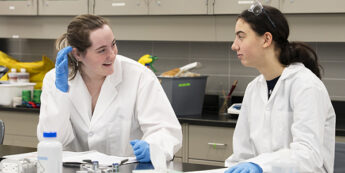 Two students in lab coats and blue gloves converse at a laboratory table with notebooks and lab equipment