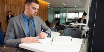 Male student in business suit and tie takes notes from textbook in brightly lit study room