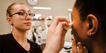 Female student applies foundation with a brush under the eye of a fellow student.