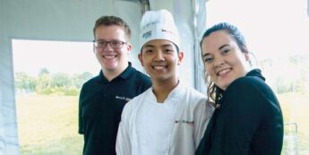 Three students, one in chef whites and two in black polos work an event for Niagara College outside in a white tent.