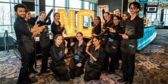 A group of students pose together at a gala event, pointing toward large illuminated NC letters.