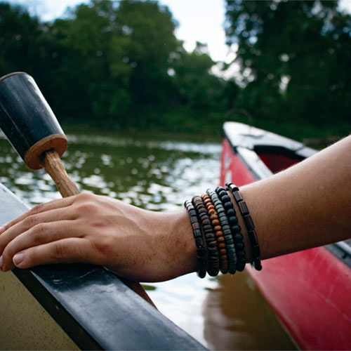 Person wearing beaded bracelets holding a paddle in a canoe next to another red canoe on a river.
