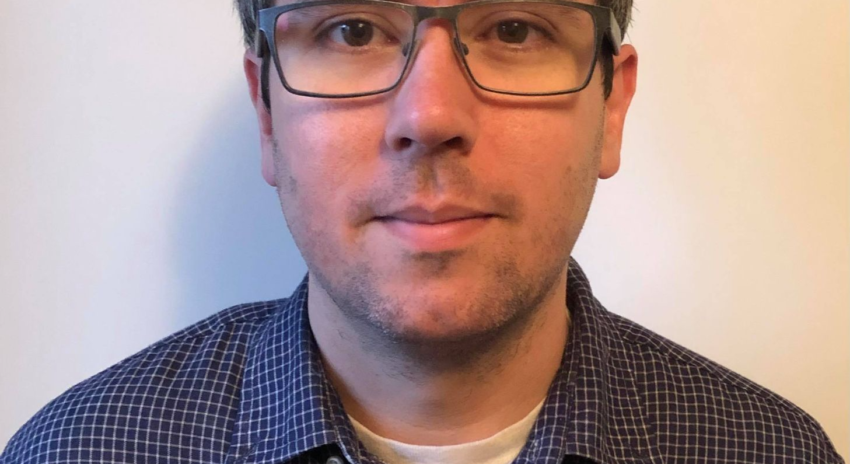 A man in glasses and a blue shirt looks at the camera and is half-smiling.