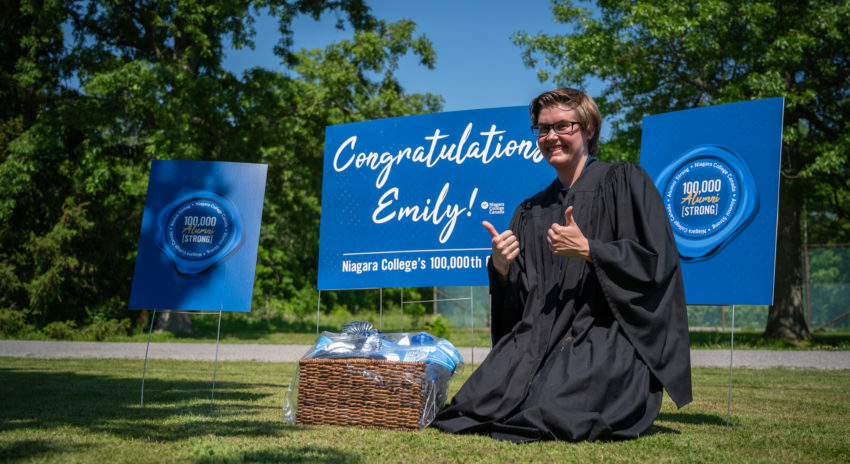 A woman wearing a graduation gown poses with the thumbs up in front of a sign announcing her as the 100,000 graduate