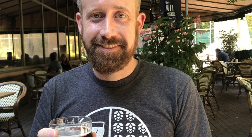 A man in a logoed t-shirt holds a beer glass and smiles.