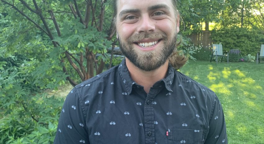 A man with a beard and his hair pulled back wears a blue button-up short-sleeve shirt and smiles for the camera.