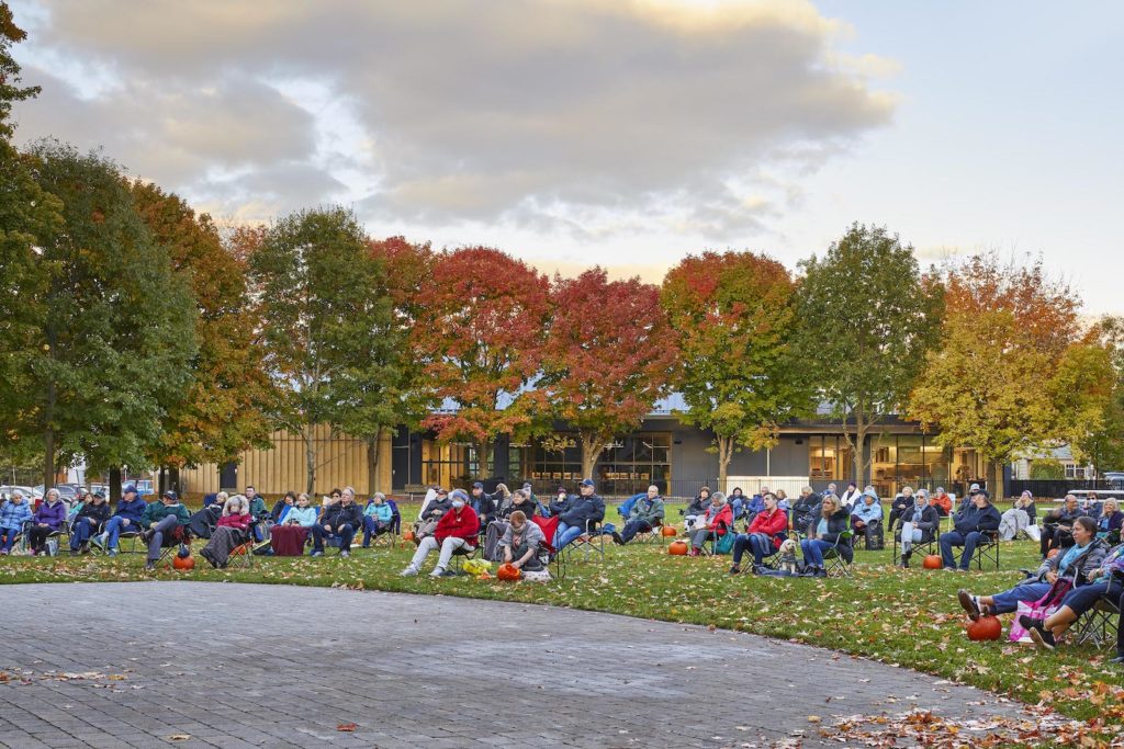 People sit outside under trees changing colours in the fall weather. They are seated on grass in park space near a modern-looking building.