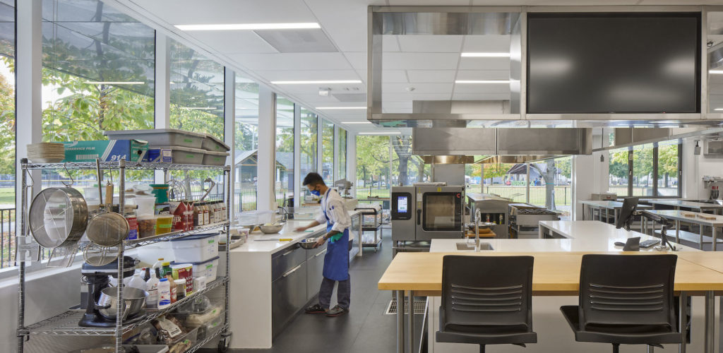 A man works in a shiny and bright, modern teaching kitchen, with shelves of ingredients and tables and chairs for students to watch.