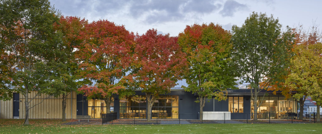A photo of the exterior of a modern one-storey building with lots of windows overlooking a park in the fall with colourful trees.