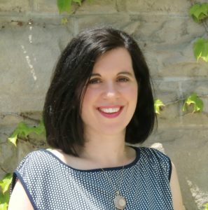 A headshot of a woman with dark, shoulder-length hair, standing agains a stone wall. 