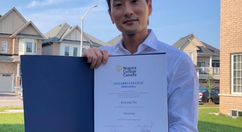 A student is pictured outside holding a diploma. He is wearing beige pants, a belt, and a light blue collared shirt.