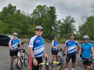 a group of cyclists stand together in a gravel parking lot