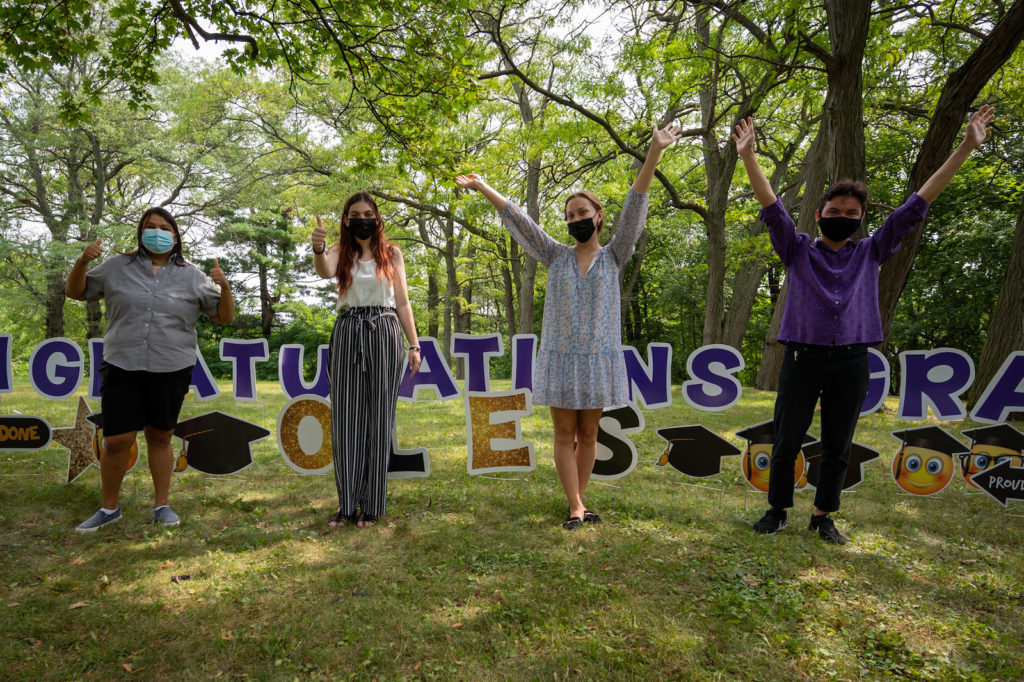 A group of four students stand in front of a congratulations sign in celebratory poses.