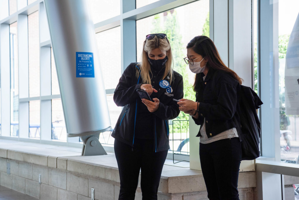 A woman shows something to another woman on her cell phone. They are standing inside a space with lots of windows.