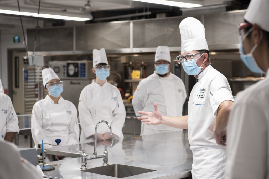 Students dressed in chefs' whites listen to their chef-professor speak in a teaching kitchen.
