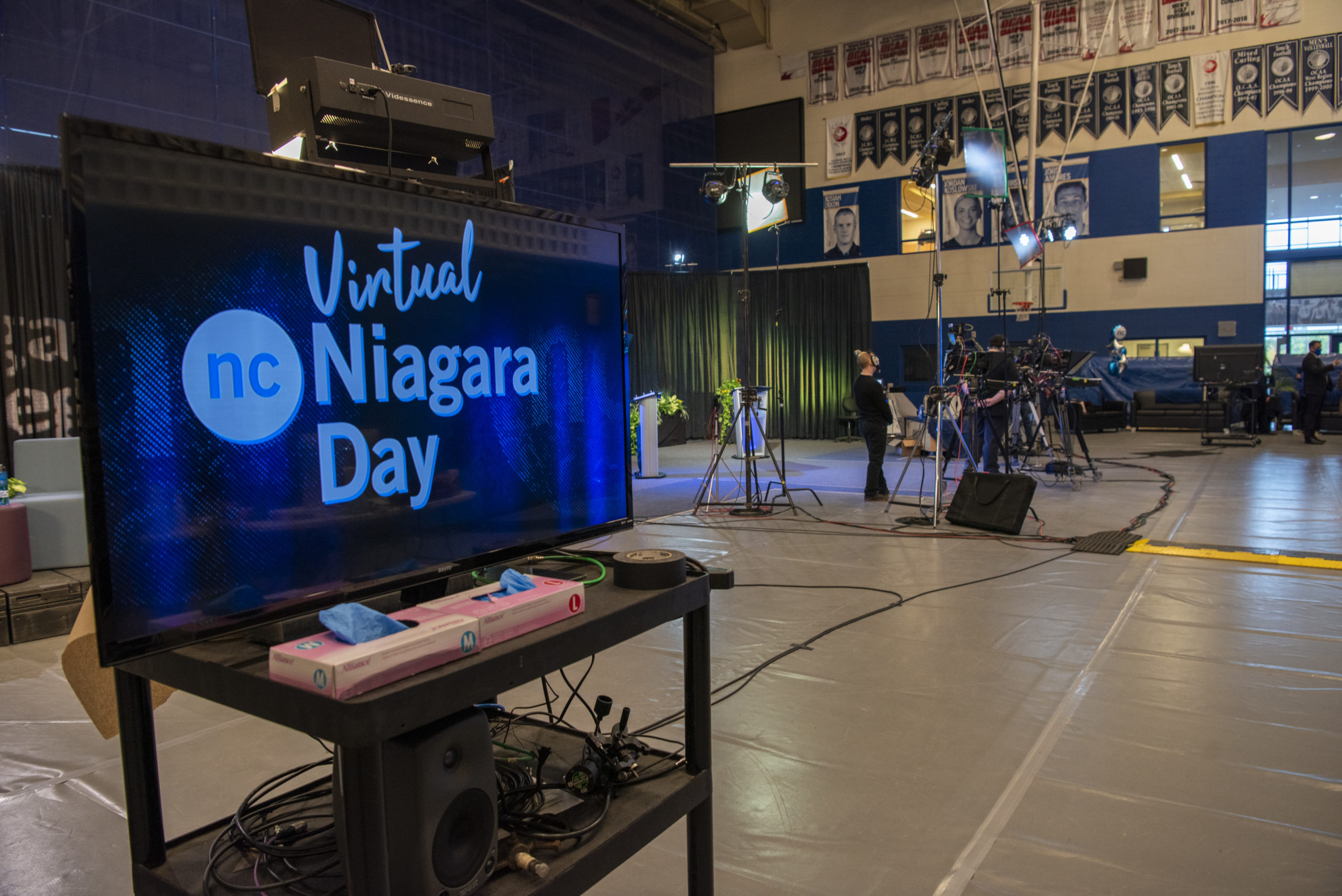 A television screen sits atop a table with the NC logo and "Niagara Day" text on a blue screen. In the background is the gym at the Welland Campus where production of the live event is taking place.