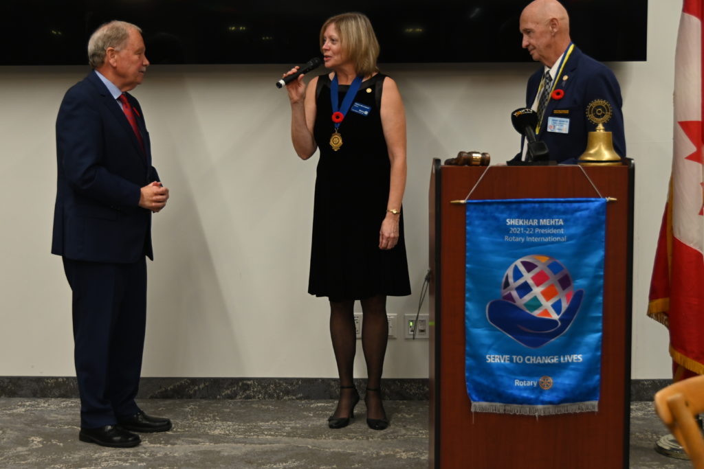 a woman in a black dress holds a microphone speaking to a man to her right, a second man stands to her left