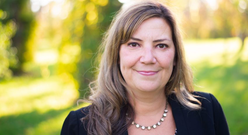 a woman with long hair wearing a black blazer smiles at the camera, a green field with the sun shining in the background