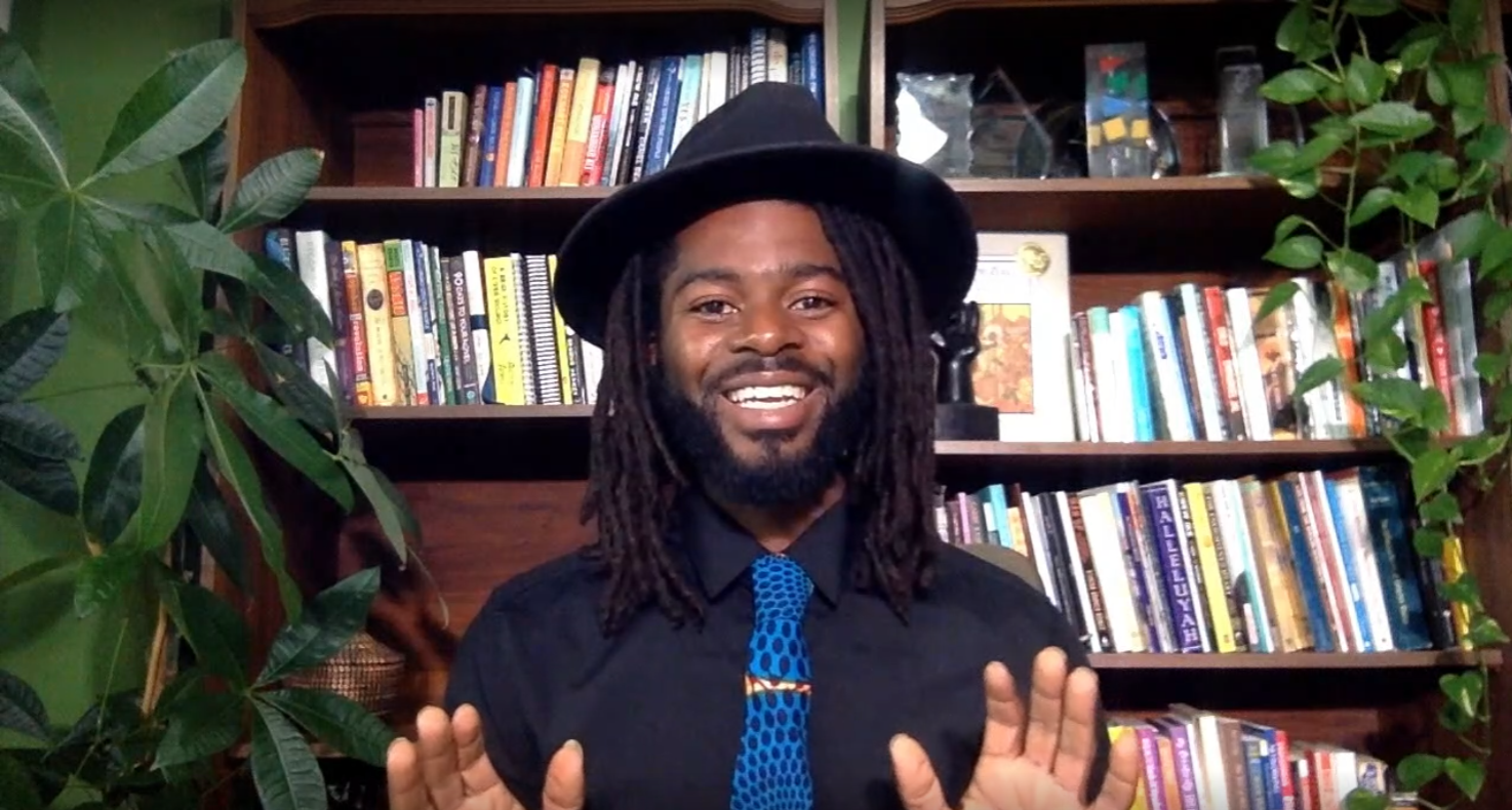 A man wearing a black hat is smiling. He is sitting in front of a bookshelf with tall plants on either side.