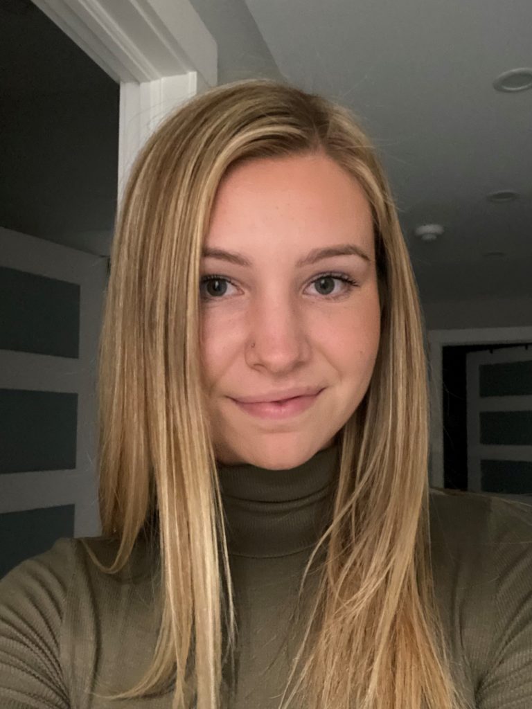 a student with long blonde hair wearing a brown turtle neck smiles at the camera