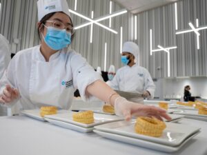 a female culinary student wearing a white outfit places a food item on a plate